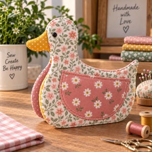 May include: A fabric duck-shaped pincushion featuring a floral pattern with pink flowers and green leaves on a white background. The duck has a yellow polka-dot beak, a black eye, and a pink wing patch decorated with a white daisy pattern.