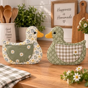 May include: Two fabric duck-shaped pot holders. The left duck features a blue and yellow floral pattern with a solid green wing pocket. The right duck has a green botanical print with a beige and white gingham wing pocket and a single daisy.