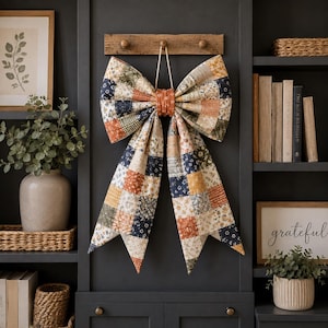 Op de afbeelding: Een grote decoratieve strik van patchworkstof met een verscheidenheid aan bloemmotieven en geometrische patronen in tinten marineblauw, gebrand oranje, crème en olijfgroen. De strik hangt aan een houten haak aan een dun wit koord.