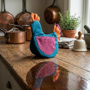 May include: A fabric chicken-shaped tea cozy in teal with an orange comb and tail feathers. It features a pink wing patch with a white leaf pattern. The item is constructed from soft textile materials and is designed to sit upright on a flat surface.