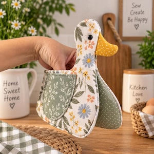 Könnte beinhalten: Ein entenförmiger Ofenhandschuh aus Stoff mit weißem Grund und einem Blumenmuster in Blau und Gelb, einem gelben Schnabel mit Punkten und einer salbeigrünen Tasche mit weißem Blattmuster. Der Handschuh hat ein schwarzes Auge und ein grün gepunktetes Futter.