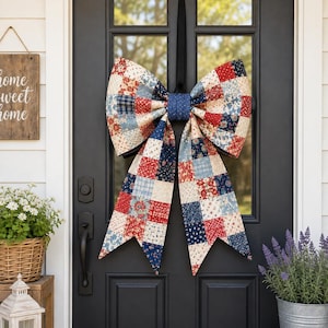 Op de afbeelding: Een grote decoratieve strik van stof met een patchworkpatroon van vierkanten in rood, marineblauw, crème en lichtblauw met diverse bloemen- en geometrische prints, in het midden voorzien van een knoop van gestructureerde marineblauwe stof.