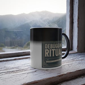 May include: A ceramic mug with a black-to-white gradient finish featuring the text DEBUGGING RITUAL in a bold, rectangular frame. The mug has a black handle and emits a thin wisp of steam from the top, indicating a hot beverage inside the container.