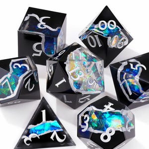 May include: A set of seven polyhedral dice featuring a black exterior with a jagged, transparent center revealing iridescent blue and green foil flakes. Each die has white engraved numbers, including a d20, d12, d10, d8, d6, d4, and a percentile die.