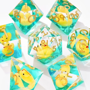 May include: A set of seven polyhedral dice made of clear teal resin, each containing a miniature yellow rubber duck inside. The dice feature gold-coloured numbers etched into the surfaces, including a d20, d12, d10, d8, d6, d4, and a percentile die.