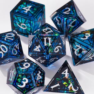 Peut inclure: Un ensemble de six dés polyédriques présentant un cadre de style vitrail bleu foncé et noir avec des inserts en feuille iridescente bleu et vert chatoyant. Chaque dé possède des chiffres argentés nets gravés sur les faces pour une visibilité claire.