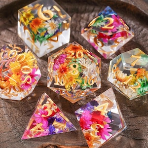 May include: A set of six polyhedral dice made of clear resin, each containing colorful dried flowers and featuring gold-colored numbers. The collection includes a d20, d12, d10, d8, d6, and d4, all displaying vibrant floral arrangements inside.