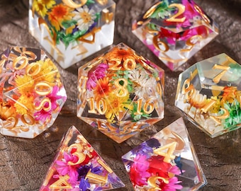 Puede incluir: Un juego de seis dados poliédricos hechos de resina transparente, cada uno con flores secas coloridas y números de color dorado. La colección incluye un d20, d12, d10, d8, d6 y d4, todos con vibrantes arreglos florales en su interior.