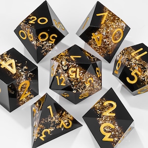 May include: A set of seven polyhedral dice made of black resin with embedded gold-colored metallic flakes. Each die features clear, recessed numbers painted in a bright gold color, including a d20, d12, d10, d8, d6, d4, and a percentile die.