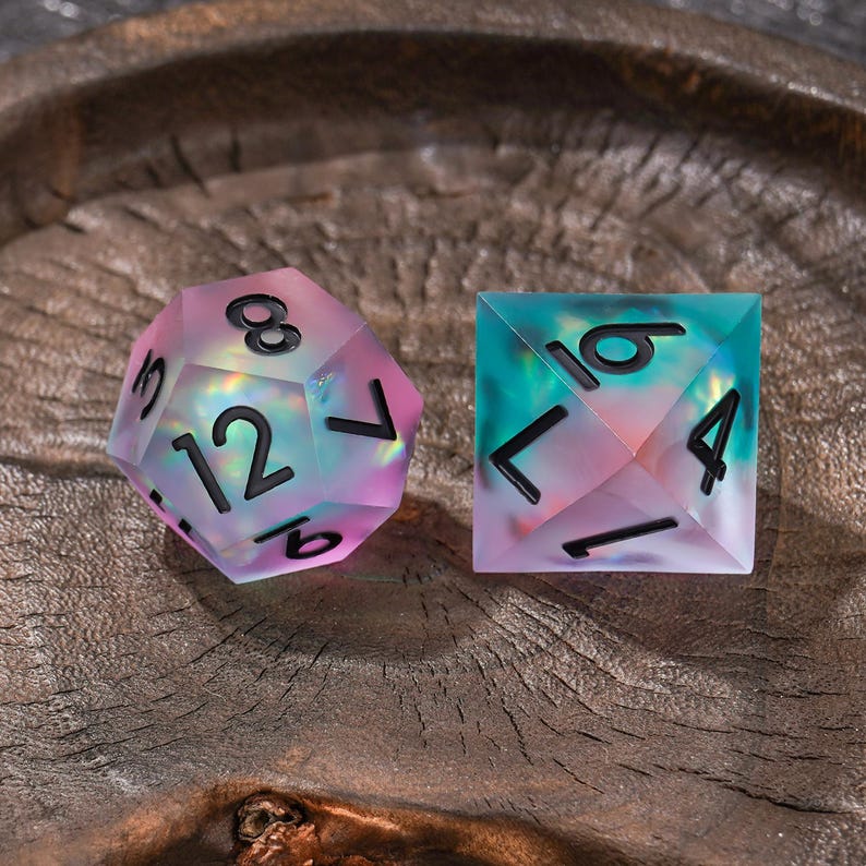 Puede incluir: Dos dados poli&eacute;dricos con un acabado transl&uacute;cido e iridiscente que presenta una mezcla de tonos rosa, azul y verde. Los dados tienen bordes afilados y est&aacute;n marcados con n&uacute;meros negros en negrita y sin remates. Uno es de veinte caras, el otro de cuatro.