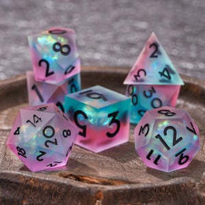 Puede incluir: Un juego de dados poli&eacute;dricos con un acabado esmerilado y semitransl&uacute;cido que presenta un degradado de tonos rosa, azul y morado. Cada dado contiene inclusiones iridiscentes brillantes y est&aacute; marcado con n&uacute;meros negros claros y en negrita.