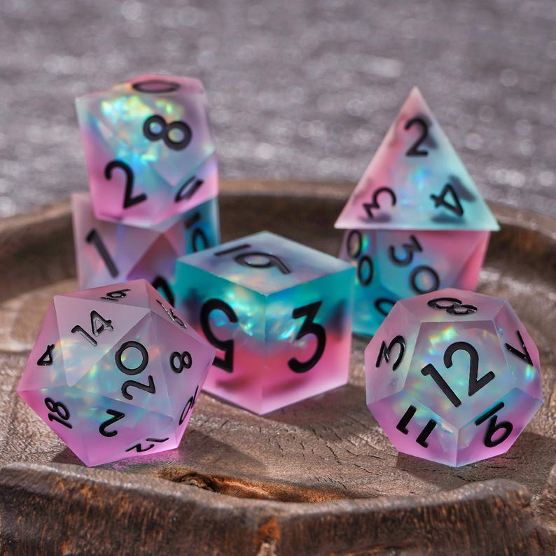 Puede incluir: Un juego de dados poli&eacute;dricos con un acabado esmerilado y semitransl&uacute;cido que presenta un degradado de tonos rosa, azul y morado. Cada dado contiene inclusiones iridiscentes brillantes y est&aacute; marcado con n&uacute;meros negros claros y en negrita.