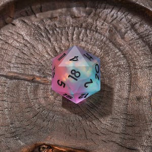 Puede incluir: Un dado de veinte caras con un degradado transl&uacute;cido de tonos rosa, morado y azul con un interior iridiscente y nublado. El dado tiene n&uacute;meros grabados en negro, incluido un dieciocho visible en la cara triangular superior.