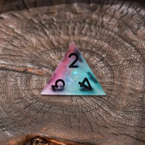 Puede incluir: Un dado triangular de cuatro caras que presenta una mezcla transl&uacute;cida y arremolinada de colores rosa, morado y verde azulado. El dado tiene los n&uacute;meros negros 2, 3 y 4 impresos en su superficie plana, con un acabado liso y pulido.