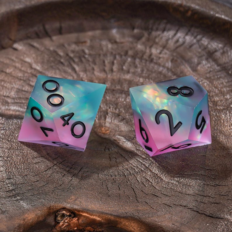Puede incluir: Dos dados poli&eacute;dricos con un degradado transl&uacute;cido de color verde azulado y rosa con un acabado iridiscente y brillante. Los dados tienen bordes afilados y n&uacute;meros grabados en negro, incluyendo un d10 que muestra 40 y 10, y un d8 que muestra 8, 2, 6 y 5.