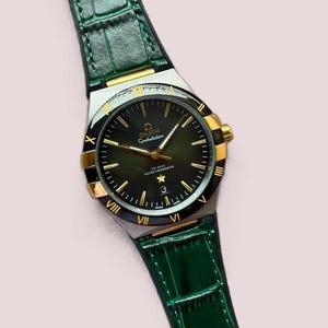 May include: An Omega Constellation wristwatch with a dark green dial, gold-tone hands, and hour markers. The bezel features gold Roman numerals. It has a stainless steel case and a textured dark green leather strap with a crocodile-like pattern.