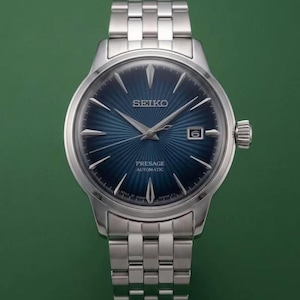 May include: A silver-tone stainless steel Seiko Presage automatic wristwatch featuring a deep blue sunray dial with a radial pattern, silver-tone hands and indices, a date window at the three o'clock position, and a multi-link metal bracelet.