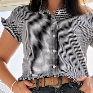 May include: A black and white gingham button-down shirt with short sleeves, a collar, and a ruffled hemline. The shirt is paired with a tan leather belt featuring a decorative metal buckle and dark gray denim jeans, creating a casual ensemble.
