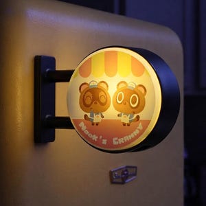 Puede incluir: Un letrero circular iluminado montado en la pared con un marco negro. La cara presenta un gráfico de dos personajes de mapache marrones con gorras y delantales bajo un toldo de rayas amarillas y blancas, con el texto Nook's Granny impreso.