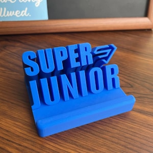 May include: A bright blue 3D printed phone stand featuring the raised text SUPER JUNIOR in a bold, blocky font. The stand has a horizontal ledge at the base for supporting a mobile device, with the band name displayed prominently on the back support.