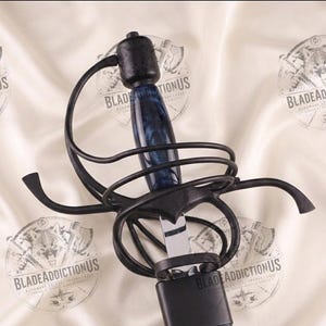 Op de afbeelding: Een rapier met een gepolijst stalen lemmet, een donkerblauw gemarmerd handvat en een complexe zwarte metalen stootplaat. Het zwaard zit gedeeltelijk in een donkerblauwe schede. De tekst BLADEADDICTIONUS is zichtbaar op de achtergrond.