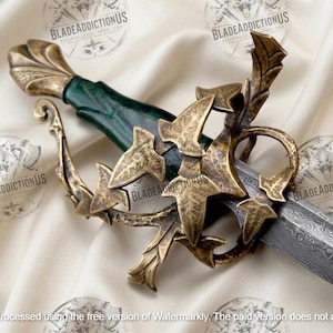 May include: A sword featuring a dark green handle and a brass-toned metal hilt designed with intricate leaf-shaped accents. The blade displays a distinct wavy Damascus steel pattern, extending from the ornate guard toward the pointed tip.