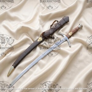 May include: A silver-toned metal sword with a wooden handle and a brass pommel, featuring a crossguard and an engraved blade. Beside the sword lies a dark brown leather scabbard with brass accents, including a pointed tip and a decorative strap.