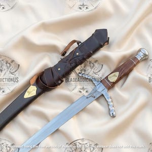 May include: A silver-toned sword with a wooden handle and a metallic pommel, featuring a gold-colored inlay on the grip. The sword is accompanied by a dark brown leather sheath with a matching gold-colored inlay and a leather strap attachment.