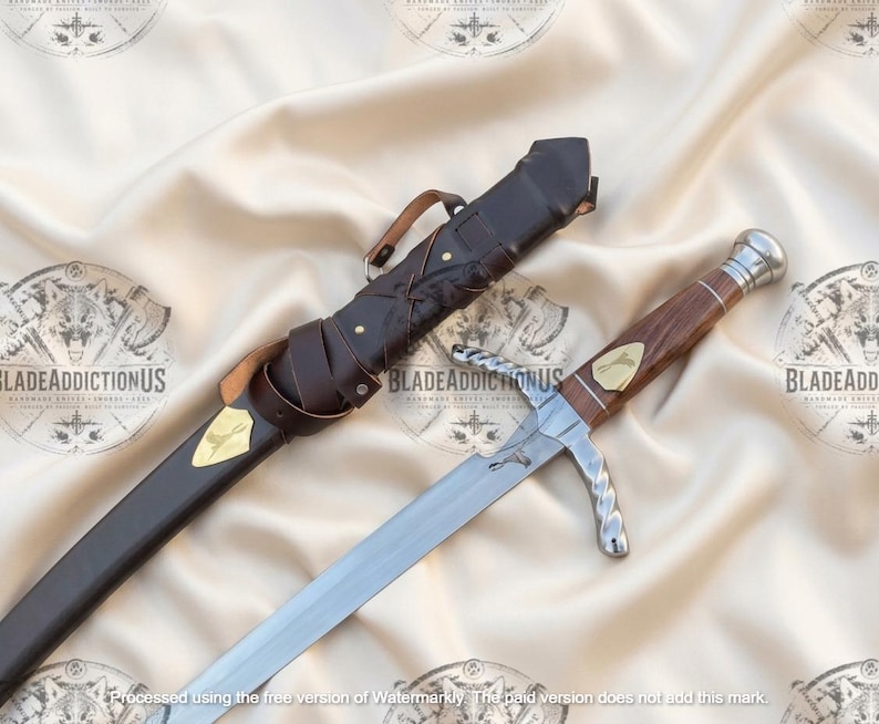 May include: A silver-toned sword with a wooden handle and a metallic pommel, featuring a gold-colored inlay on the grip. The sword is accompanied by a dark brown leather sheath with a matching gold-colored inlay and a leather strap attachment.