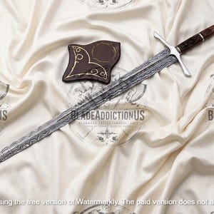 May include: A long sword with a patterned Damascus steel blade, a silver crossguard, and a dark brown wooden handle with silver accents. A small, dark brown, shield-shaped leather sheath with gold-colored decorative line work is placed nearby.