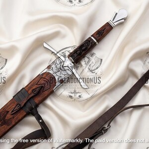 May include: A sword with a dark brown wooden handle and scabbard featuring intricate black engravings. The hilt has a silver-colored pommel and crossguard. A dark brown leather strap is attached to the scabbard. The text BLADEADDICTIONUS is visible.