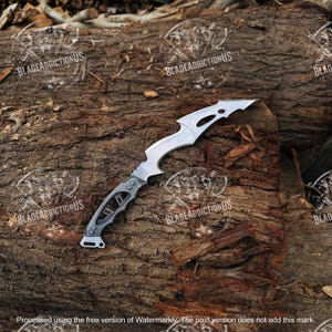 May include: A silver-toned metal knife featuring a curved, serrated blade with a pointed tip and a skeletal handle design with cutouts. The blade has a distinct notched spine and a metallic finish, showcasing a complex, angular construction throughout.