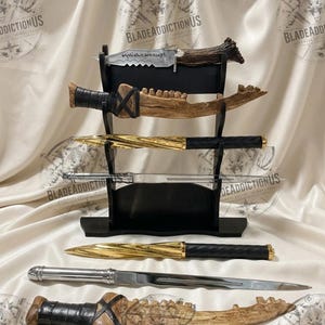 May include: A black tiered display stand holding three knives, with three additional knives placed horizontally in front. The collection includes a serrated blade with etched text, two bone-textured knives with black wraps, two gold-toned spiral knives, and a silver-toned blade.