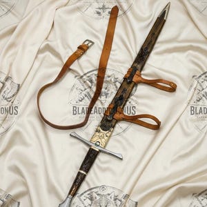 May include: A sword with a dark wood handle featuring intricate carvings, a brass crossguard, and a matching wooden scabbard with brass accents. A tan leather belt with a metal buckle is attached to the scabbard, which has two leather straps for carrying.