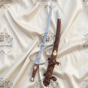 May include: A curved steel sword with a wooden handle and a silver-toned crossguard. The sword features a small gold-toned inlay on the grip. Beside it lies a brown leather scabbard with silver-toned metal fittings and leather straps.