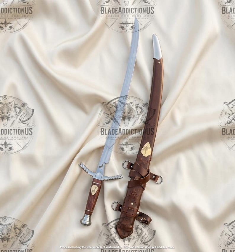 May include: A curved steel sword with a wooden handle and a silver-toned crossguard. The sword features a small gold-toned inlay on the grip. Beside it lies a brown leather scabbard with silver-toned metal fittings and leather straps.