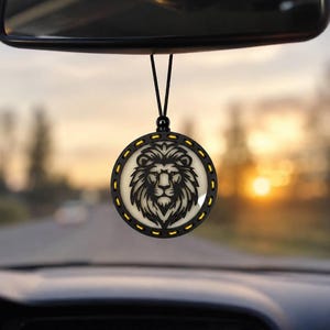May include: A circular car air freshener hanging from a rearview mirror, featuring a black frame with yellow accents and a detailed black lion head silhouette centered on a light-colored background, suspended by a thin black cord.