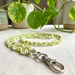 May include: A lanyard crafted from small, round beads in alternating shades of lime green and cream, arranged in a repeating floral pattern. The beaded cord is finished with silver-toned metal crimp beads and a sturdy silver-toned lobster clasp.