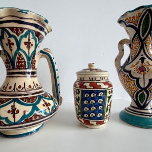 Puede incluir: Dos jarrones de cerámica con bordes ondulados y asas laterales, y un pequeño tarro con tapa. Las tres piezas presentan patrones geométricos y florales pintados a mano en tonos azul verdoso, azul oscuro, rojo y amarillo sobre un fondo crema.