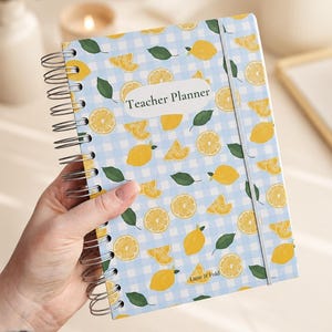 May include: A spiral-bound teacher planner featuring a light blue and white gingham pattern decorated with yellow lemon slices and green leaves. The cover includes a white label with the text Teacher Planner and the brand name Lune & Fold.