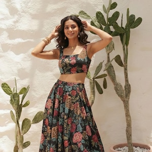 Könnte beinhalten: Ein zweiteiliges Outfit bestehend aus einem ärmellosen bauchfreien Oberteil mit quadratischem Ausschnitt und einem bodenlangen Rock mit hoher Taille. Beide Teile haben einen schwarzen Grundstoff mit einem bunten Blumenmuster in Rot, Blau und Gold.