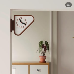 Op de afbeelding: Een dubbelzijdige wandklok met een bruin driehoekig frame en een witte wijzerplaat met een ruitjespatroon. De klok heeft zwarte cijfers van 1 tot 12 en zwarte uur- en minutenwijzers met een klein rood middelpunt op een bruine beugel.