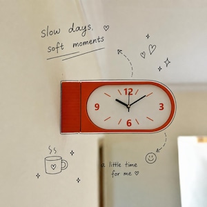 Puede incluir: Un reloj de pared naranja y blanco con forma rectangular redondeada, con manecillas negras y los números 3, 6, 9 y 12 en naranja. La pared muestra ilustraciones negras dibujadas a mano de corazones, estrellas, una taza, una cara sonriente y el texto "slow days, soft moments" y "a little time for me".