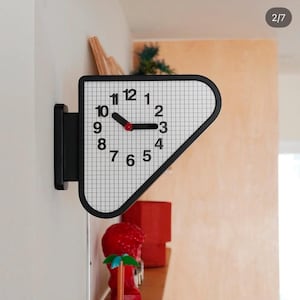 Puede incluir: Un reloj de pared con marco negro y esfera blanca con un patrón de cuadrícula negra. El reloj tiene números negros del 1 al 12, manecillas negras para las horas y los minutos, y un pequeño punto central rojo. El reloj tiene forma triangular.