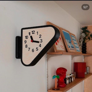 Puede incluir: Un reloj de pared de doble cara con marco negro y esfera de madera clara. El reloj presenta números arábigos negros del 1 al 12, manecillas negras para las horas y los minutos, y una pequeña tapa central roja. Montado en la pared con un soporte.