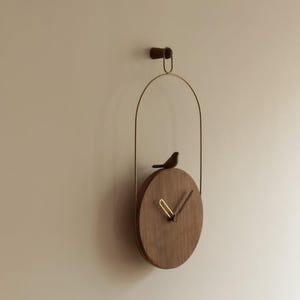 Puede incluir: Un reloj de pared circular de madera con acabado marrón oscuro, suspendido por un arco de metal dorado fino desde un soporte de pared de madera a juego. Un pequeño pájaro de madera tallada se asienta sobre la esfera, con manecillas doradas y marrones.