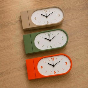 Puede incluir: Tres relojes de escritorio ovalados en beige, verde oliva y naranja brillante. Cada reloj presenta una esfera blanca con manecillas negras para las horas y los minutos, y números en las posiciones 12, 3, 6 y 9 a juego con el color del marco.