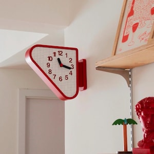 May include: A double-sided wall clock with a red frame and a white face featuring a black grid pattern. The clock has black hour and minute hands, a red center point, and black numerals from 1 to 12 arranged in a triangular shape.