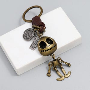 May include: A bronze-toned metal keychain featuring a skull-shaped charm with hollow eyes and a stitched mouth, attached to a brown leather cord. It includes a circular charm with concentric circles and a rectangular tag with the text, "...er about you, kes my heart, one to be free."