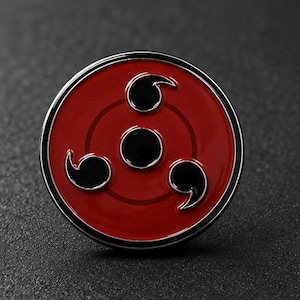 May include: A circular metal enamel pin featuring a vibrant red background with a black central circle and three black comma-shaped tomoe symbols arranged symmetrically around the center, all enclosed within a polished silver-toned metal border.